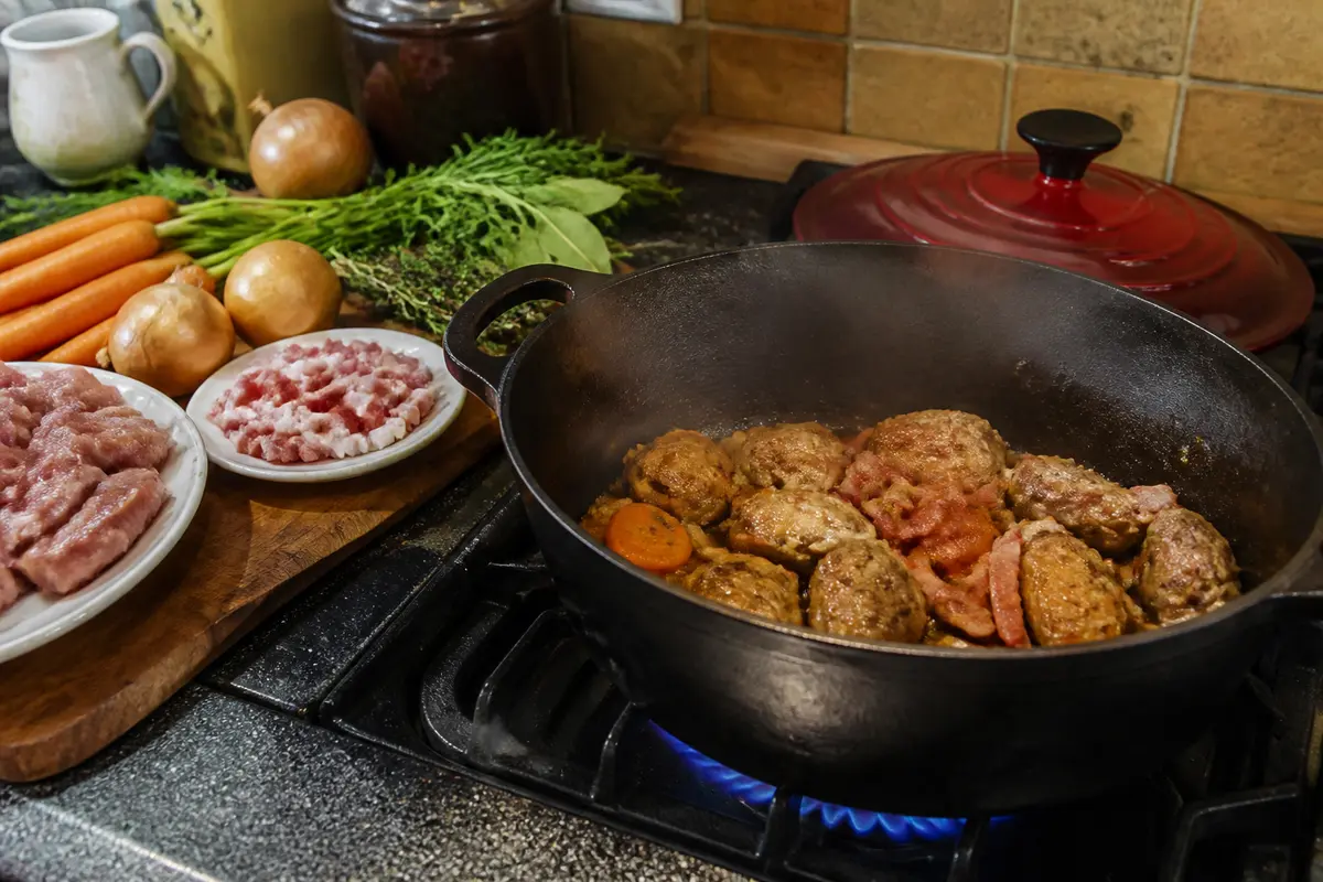 Cocotte en fonte sur gaz, cuisson de viande avec légumes et herbes fraîches à côté.